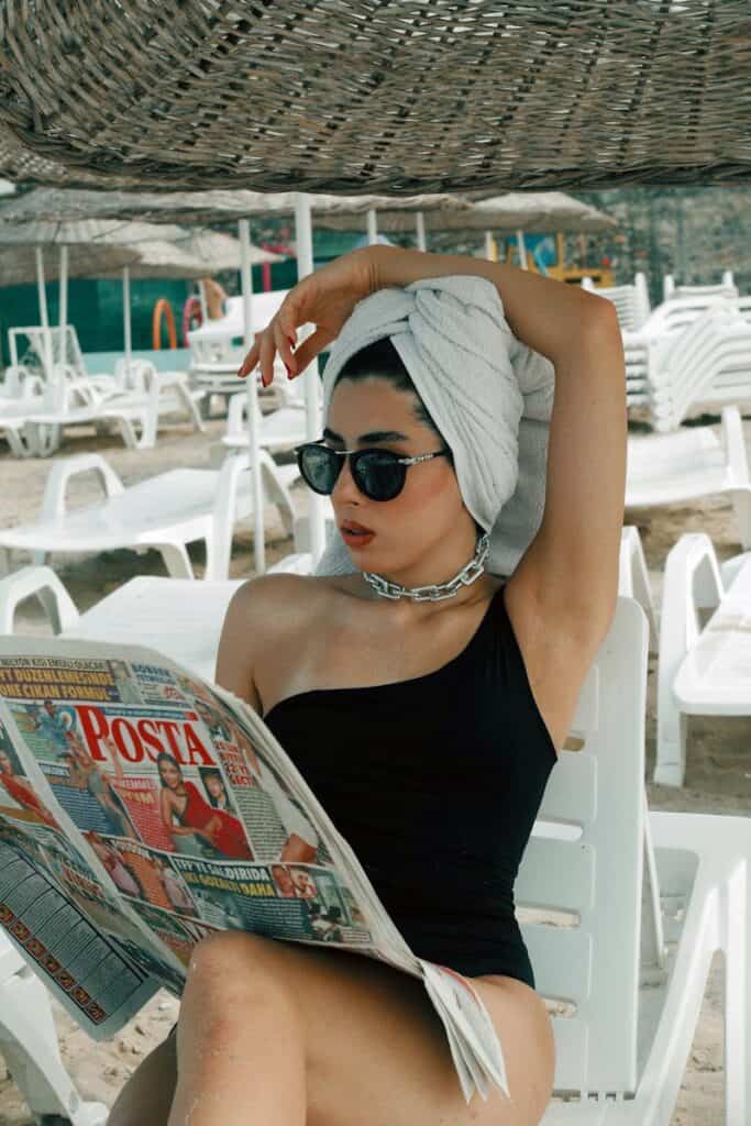 Relaxed woman sunbathing at the beach reading newspaper, wearing sunglasses and towel wrapped on head.